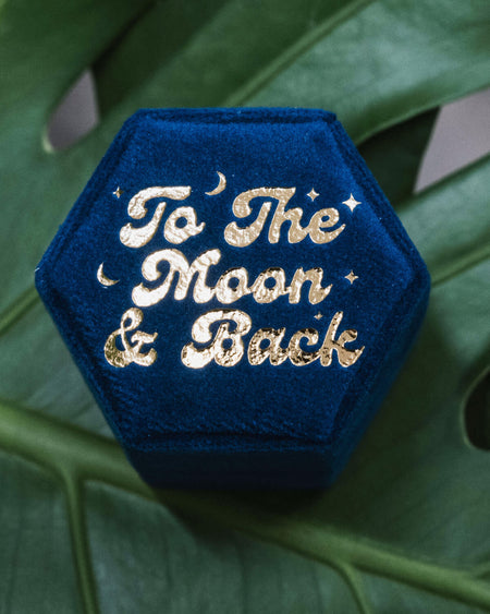 The top view of a hexagonal blue velvet ring box featuring the gold-embossed phrase “To The Moon & Back,” surrounded by moon and star illustrations, displayed on green foliage.