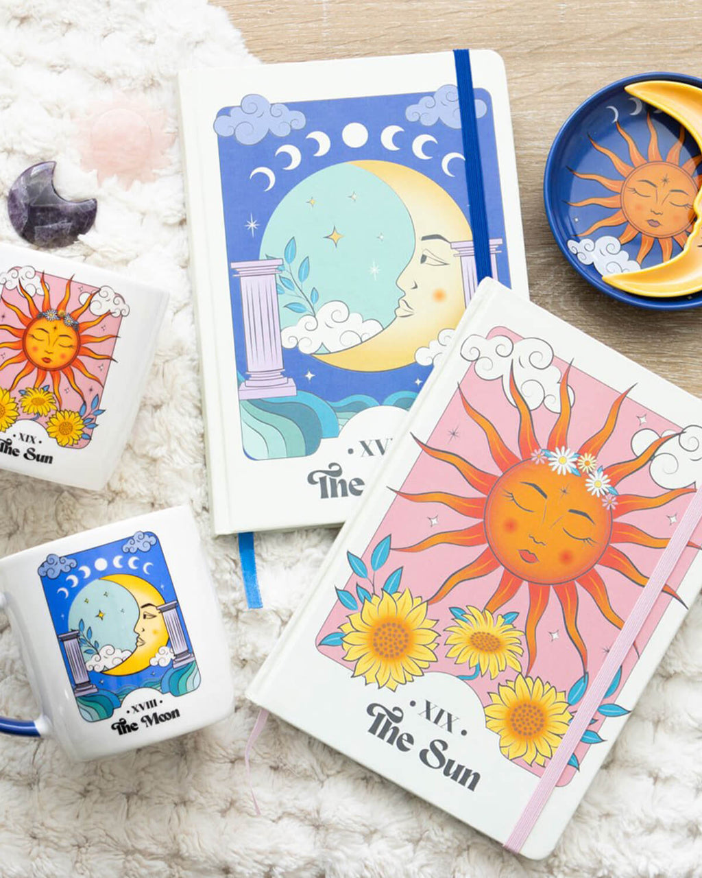 Flat lay of celestial-themed stationery set – The Sun and The Moon A5 notebooks, matching mugs, and a sun and moon trinket dish are styled on a soft, cream blanket with dried flowers, crystals, and other mystical accessories.