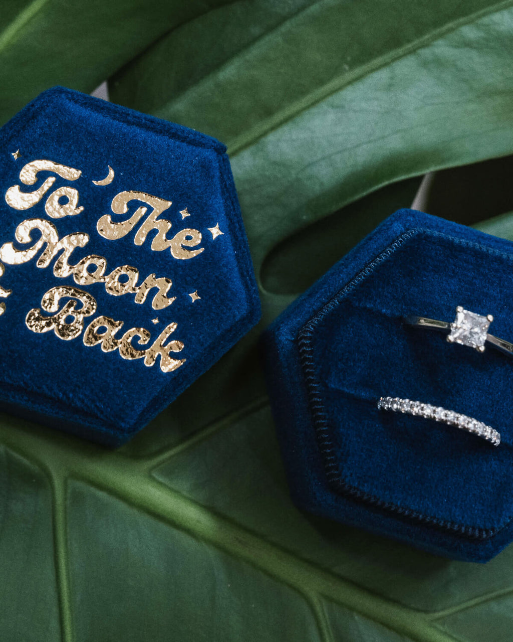 Two blue velvet hexagonal ring boxes, one showcasing gold-embossed “To The Moon & Back” and the other opened to reveal a sparkling engagement ring and wedding band, placed artistically on a green leafy surface.