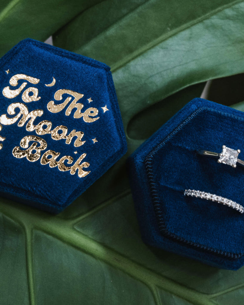 Two blue velvet hexagonal ring boxes, one showcasing gold-embossed “To The Moon & Back” and the other opened to reveal a sparkling engagement ring and wedding band, placed artistically on a green leafy surface.