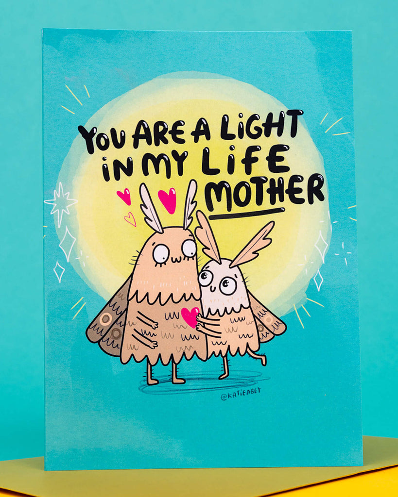 Bright and quirky A6 greeting card by Rebel Romance featuring two illustrated moth characters hugging under a glowing light with the text “You Are A Light In My Life Mother.” Designed by Katie Abey, this colourful card is perfect for Mother’s Day, birthdays, or to show appreciation. Printed in the UK on 250gsm card with a blank inside for personal messages.