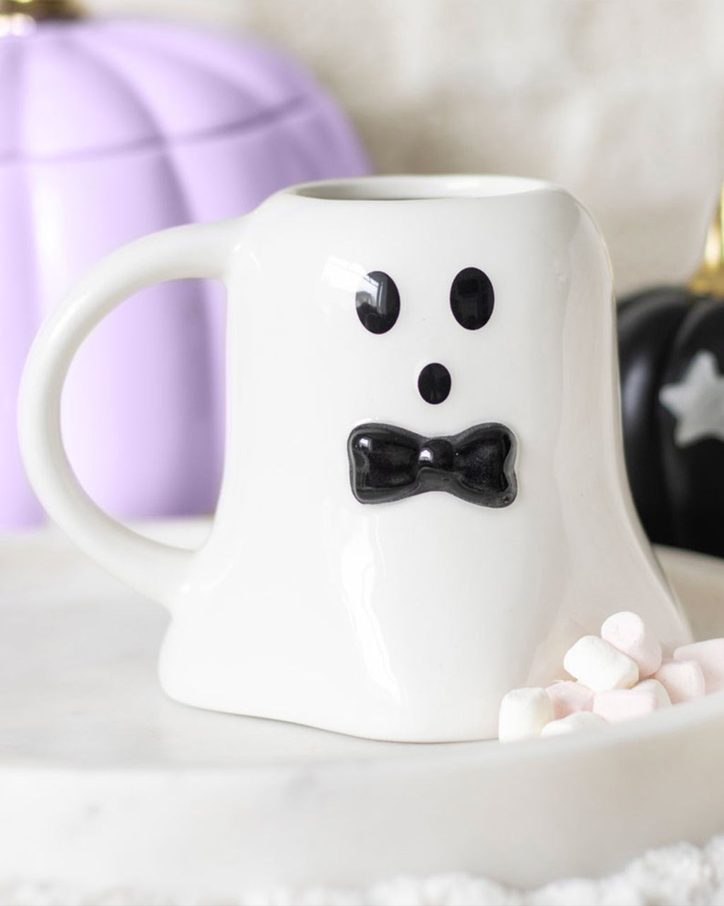 White ceramic ghost-shaped mug with a black bow tie on a round white tray, styled with mini marshmallows and Halloween pumpkins in the background.