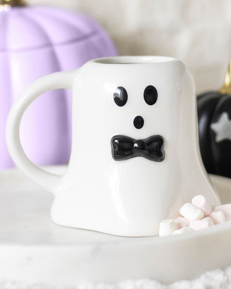 White ceramic ghost-shaped mug with a black bow tie on a round white tray, styled with mini marshmallows and Halloween pumpkins in the background.