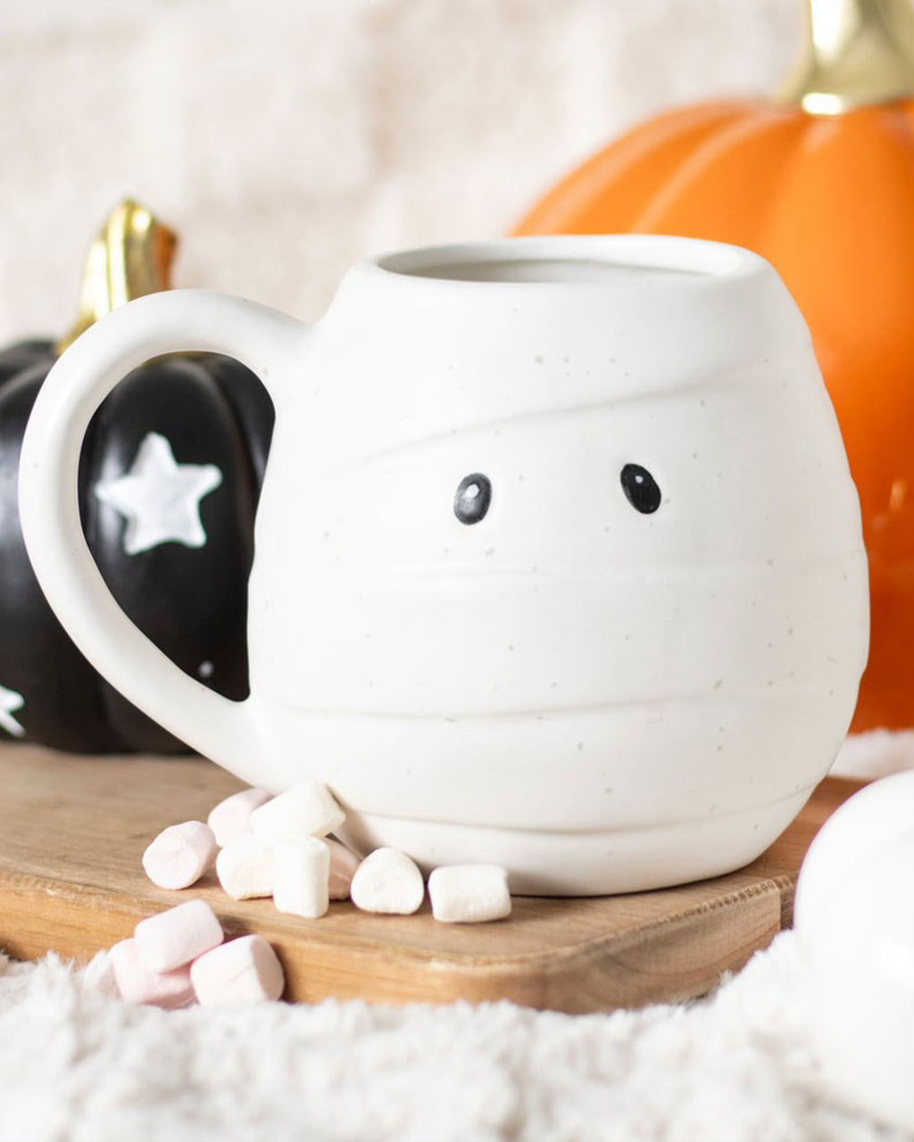 Close-up of a single mummy-shaped mug with marshmallows scattered at the base, black and orange pumpkins in the background. Ideal for Halloween hot chocolate.