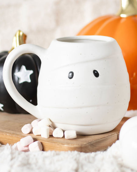 Close-up of a single mummy-shaped mug with marshmallows scattered at the base, black and orange pumpkins in the background. Ideal for Halloween hot chocolate.