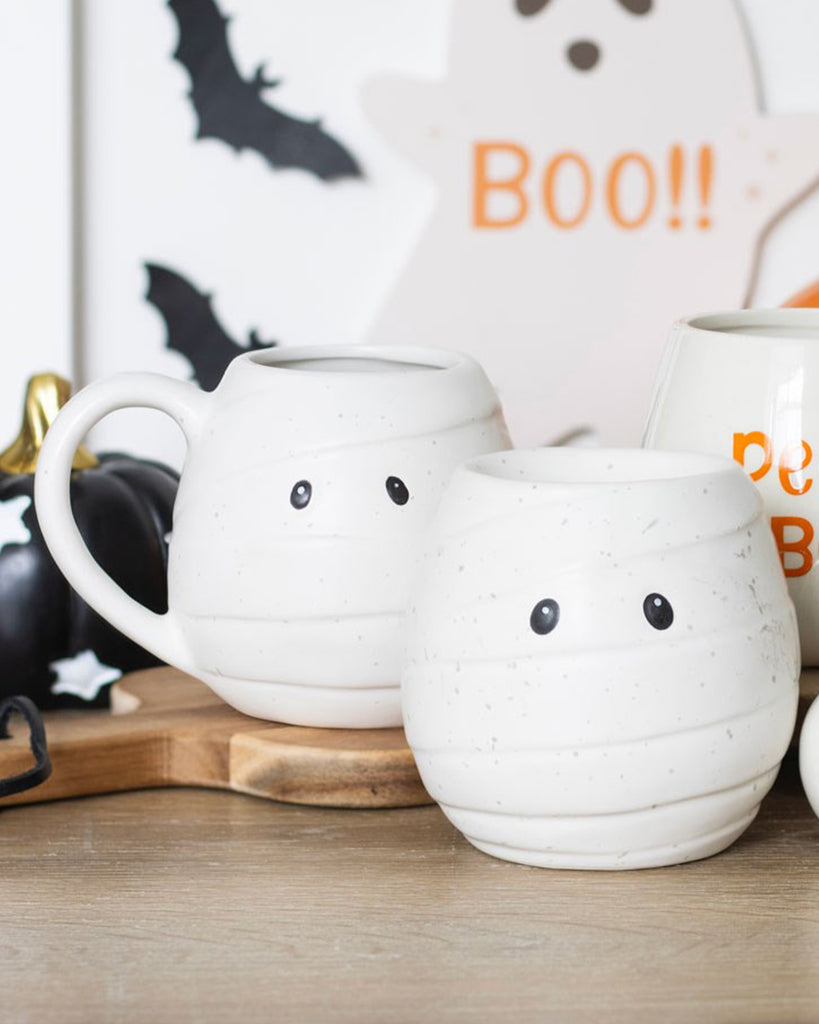 A white ceramic mummy mug and white ceramic mummy candle holder both with wide black eyes, styled with Halloween decorations including bats and ghosts. Perfect for spooky autumn drinks.