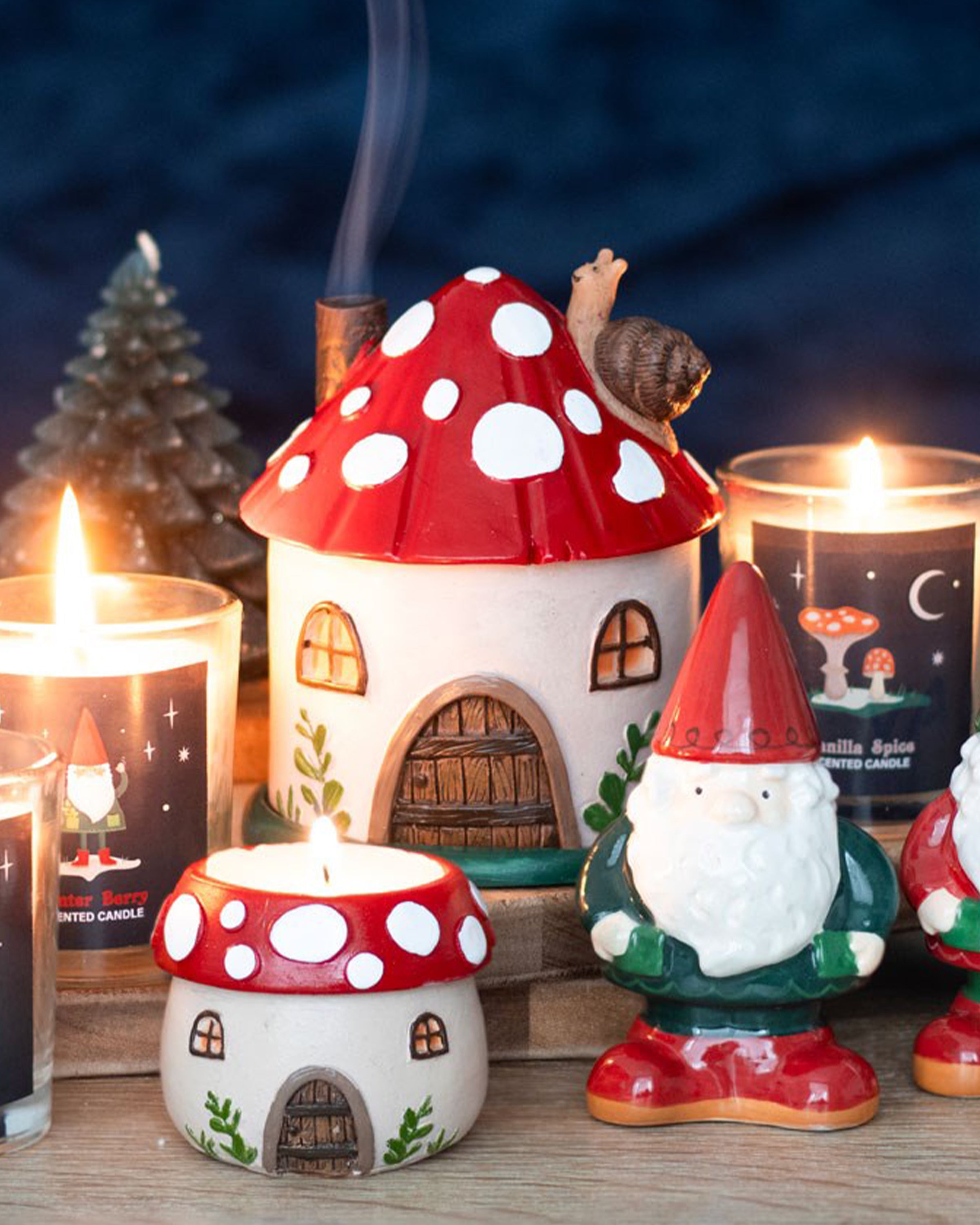 A cosy home scene featuring a red-and-white mushroom house incense burner with a snail on the roof. Lit candles and festive gnomes surround it. Smoke curls from the chimney. Product sold by Rebel Romance, a UK small business selling whimsical alternative homeware and gifts.