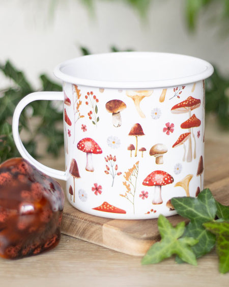 A beautifully styled shot of the mushroom enamel mug, resting on a wooden board surrounded by ivy and a decorative glass mushroom ornament. A must-have for woodland and cottagecore lovers.