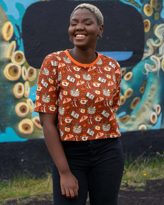 Chish smiles wearing the Rebel Romance Mushroom Tea Party cropped tee, paired with black jeans against a colourful mural wall.