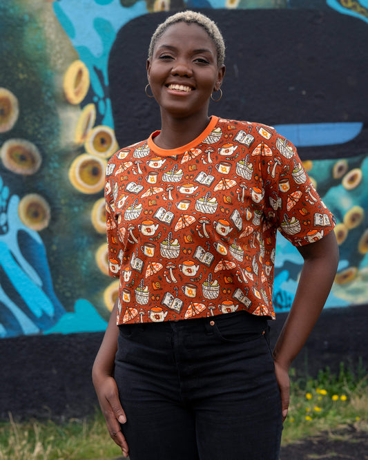 Chish smiles wearing the Rebel Romance Mushroom Tea Party cropped tee, paired with black jeans against a colourful mural wall.