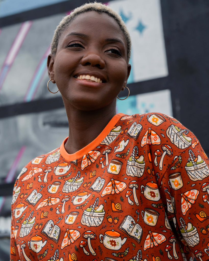 Chish poses in the Rebel Romance Mushroom Tea Party cropped tee, featuring a rustic orange background with cosy cottagecore illustrations.