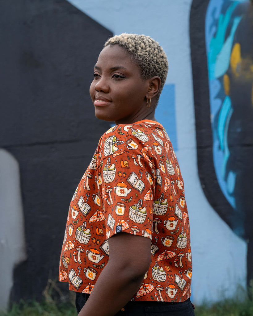 Close-up portrait of Chish smiling in the Rebel Romance Mushroom Tea Party cropped tee, showing print details of mushrooms, teacups, and moths.