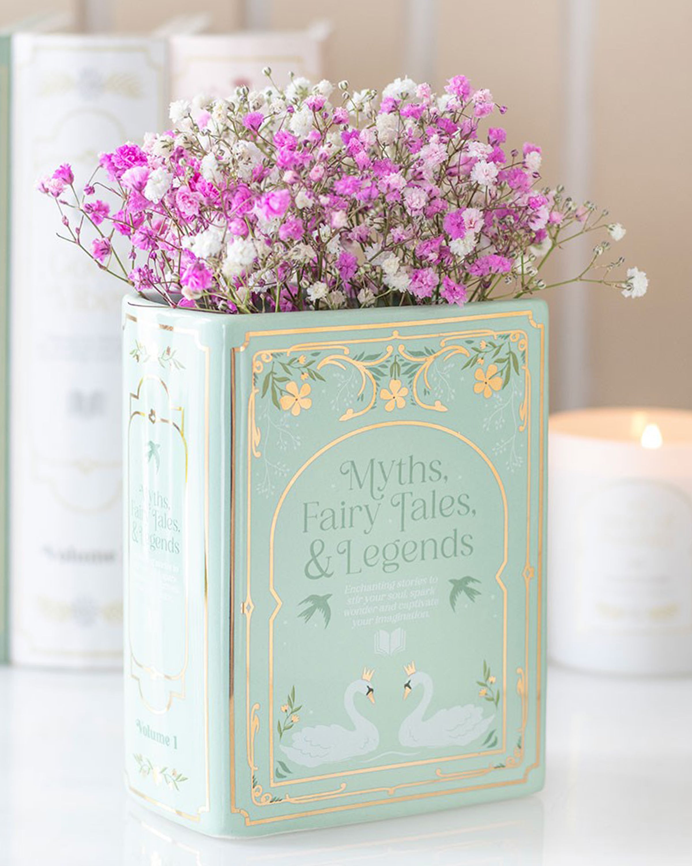 Styled display of the Rebel Romance Myths, Fairy Tales & Legends Green Book Shaped Vase filled with pink and white flowers. A ceramic vase shaped like a book, perfect for bookworms, fantasy lovers, or anyone who wants to add a whimsical literary flourish to their home.