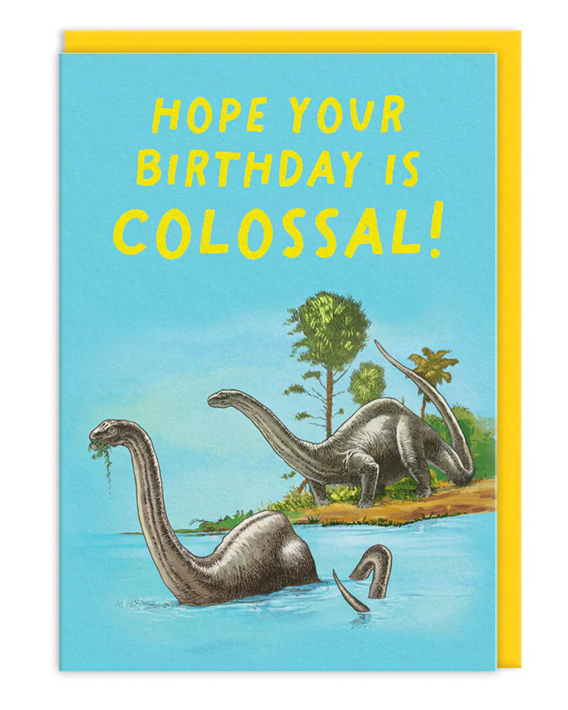 A6 dinosaur birthday card featuring illustrated brontosauruses by water with yellow text reading “Hope Your Birthday Is Colossal!” on a blue sky background, paired with a bright yellow envelope.