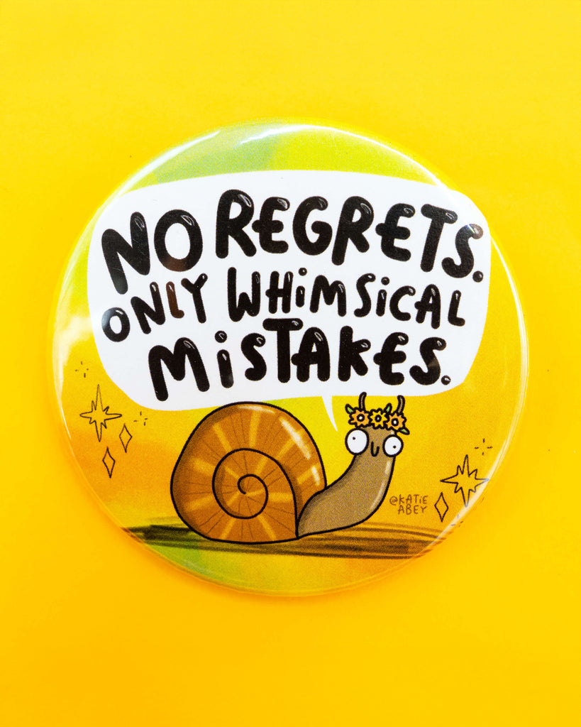 The Rebel Romance Whimsical Mistakes Snail Badge by Katie Abey photographed flat on a bright yellow background. Featuring a cartoon snail wearing a flower crown and the words “No regrets. Only whimsical mistakes.” A cheerful motivational badge from a UK brand known for alternative fashion and joyful gifts.