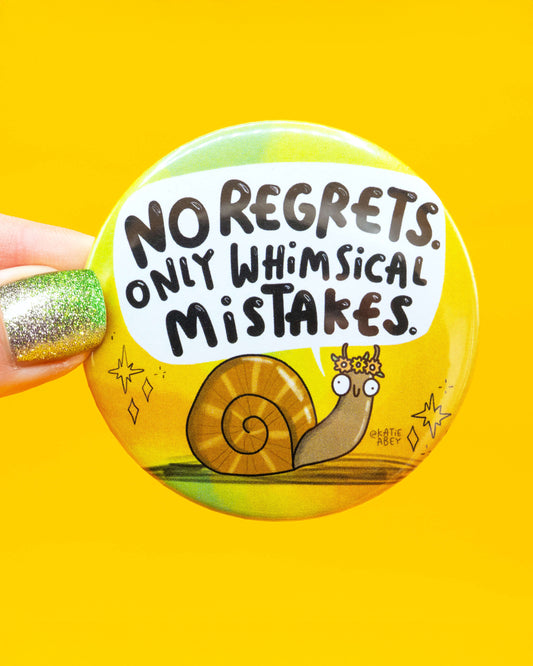 Hand holding the Rebel Romance Whimsical Mistakes Snail Badge by Katie Abey against a bold yellow background. The badge features a wide-eyed snail in a flower crown with the words “No regrets. Only whimsical mistakes.” A vibrant, quirky accessory from a UK small business that embraces millennial-friendly, feel-good designs.