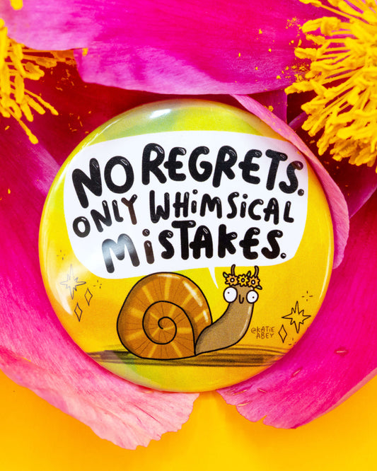 Close-up of the Rebel Romance Whimsical Mistakes Snail Badge by Katie Abey, nestled inside bright pink flower petals. The colourful badge features a cute snail with a flower crown and the phrase “No regrets. Only whimsical mistakes.” A quirky and motivational gift idea from a small UK business that celebrates alternative fashion and aesthetics.