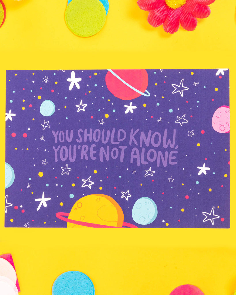 An A6 postcard featuring a colourful cosmic scene with planets, stars, and tiny scattered dots on a dark purple background. The central text reads: “You Should Know, You’re Not Alone.” A positive and comforting design by Katie Abey, printed on premium card.