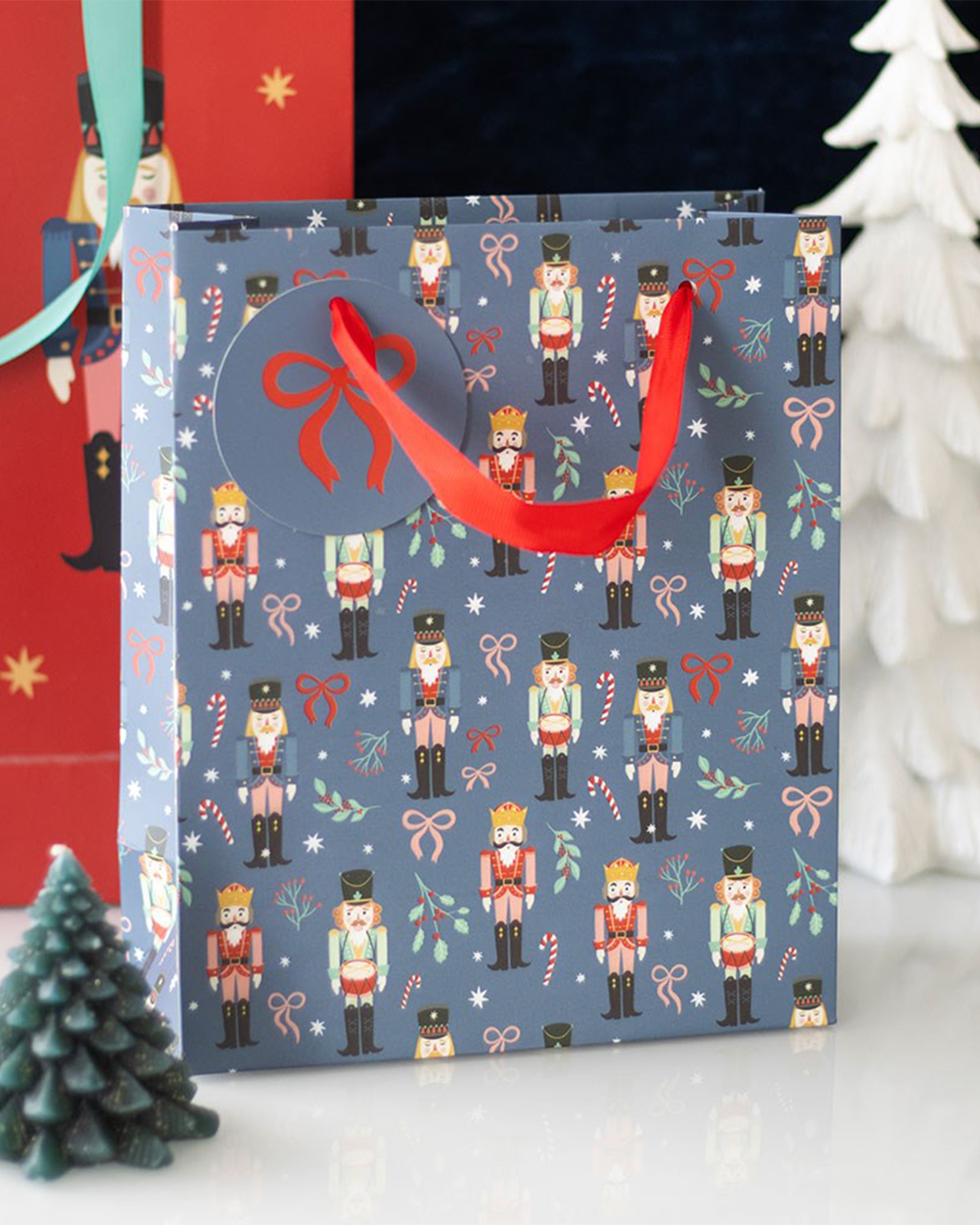 Styled photo of the Rebel Romance medium nutcracker print gift bag displayed next to Christmas trees. Featuring red ribbon handles, a festive tag, and a cheerful nutcracker, candy cane, and holly design—perfect for wrapping seasonal gifts in alternative style.