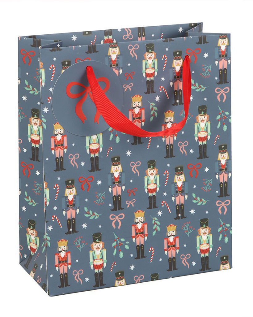 Side view of the Rebel Romance blue nutcracker gift bag showcasing its vibrant all-over Christmas design with red and pink bows, candy canes, stars and festive toy soldiers. The sturdy bag includes a gift tag and red ribbon handles for easy carrying.