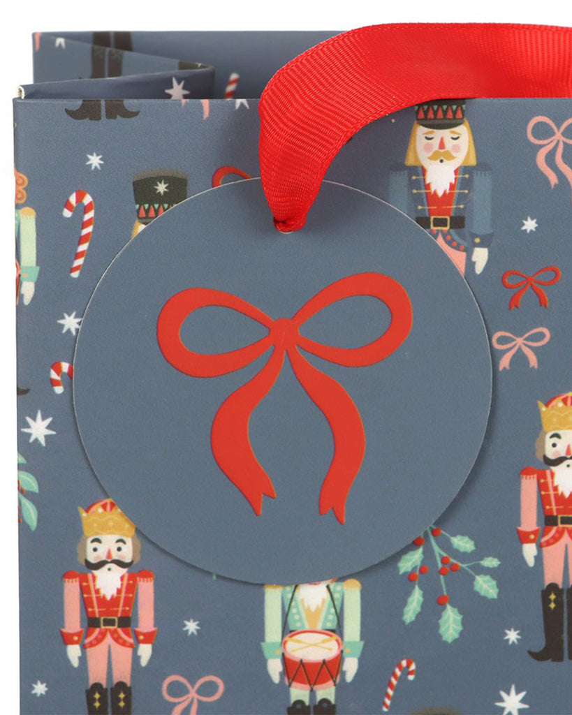 Close-up of Rebel Romance’s medium nutcracker print Christmas gift bag showing a circular gift tag with a red bow illustration and matching red ribbon handle. The festive nutcracker design features candy canes, holly sprigs, stars and bows on a blue background.