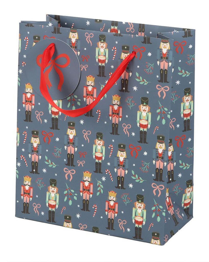 Medium blue Christmas gift bag from Rebel Romance featuring an all-over festive nutcracker pattern with candy canes, holly, stars and bows. Includes a round matching gift tag with red ribbon handles. Ideal for wrapping Christmas presents and Secret Santa surprises.