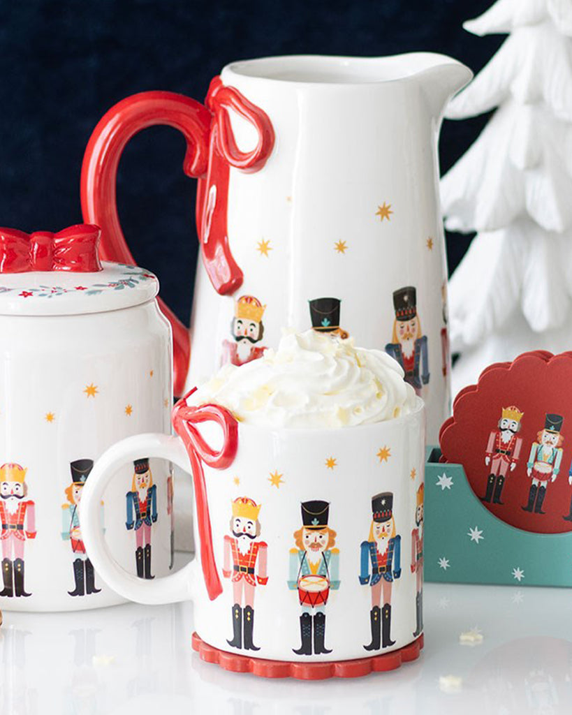 Festive ceramic mug by Rebel Romance featuring a nutcracker soldier design and red sculpted bow handle, styled with matching jug and jar in a cosy Christmas setup with whipped cream and gold chocolate coins.