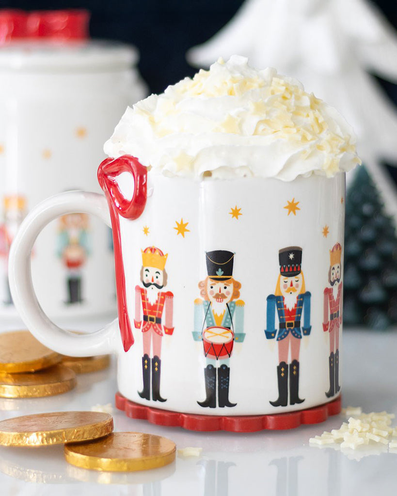 Cosy Christmas scene featuring the Rebel Romance Nutcracker Mug filled with whipped cream, surrounded by chocolate coins, festive decorations, and coordinating nutcracker-themed tableware.
