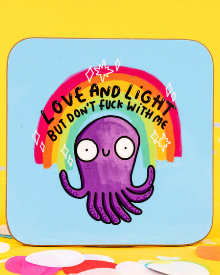 A brightly coloured square coaster featuring a cartoon purple octopus with wide eyes and arms raised. Above its head is a rainbow with the text “Love and Light But Don’t Fuck With Me.” The background is light blue, and the coaster is photographed on a yellow surface with pink confetti.