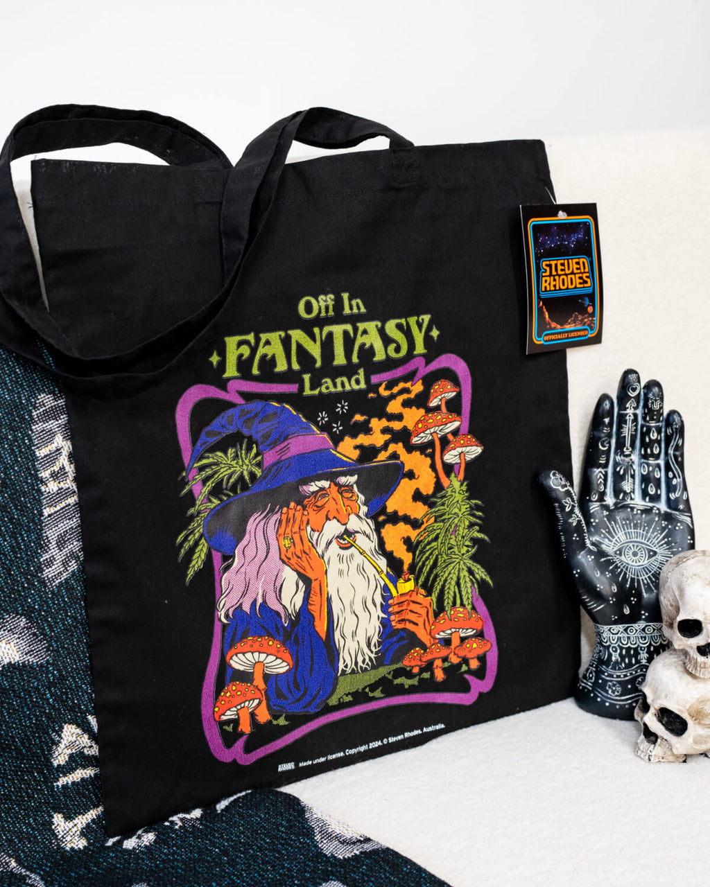 Steven Rhodes black tote bag featuring a whimsical design titled ‘Off in Fantasy Land.’ The artwork depicts a wizard sipping a drink surrounded by mushrooms and magical plants, with bold purple, orange, and green tones. The bag includes sturdy handles and an eco-friendly label, combining functionality with quirky charm.