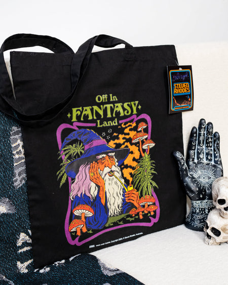 Steven Rhodes black tote bag featuring a whimsical design titled ‘Off in Fantasy Land.’ The artwork depicts a wizard sipping a drink surrounded by mushrooms and magical plants, with bold purple, orange, and green tones. The bag includes sturdy handles and an eco-friendly label, combining functionality with quirky charm.