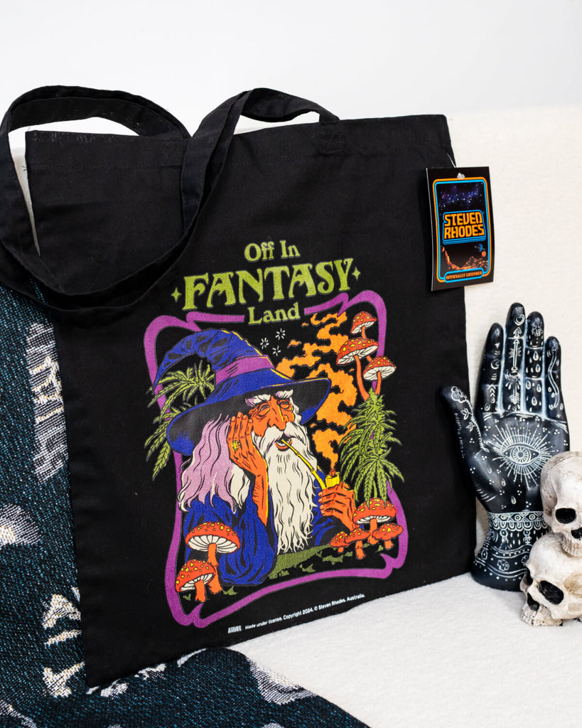Steven Rhodes black tote bag featuring a whimsical design titled ‘Off in Fantasy Land.’ The artwork depicts a wizard sipping a drink surrounded by mushrooms and magical plants, with bold purple, orange, and green tones. The bag includes sturdy handles and an eco-friendly label, combining functionality with quirky charm.