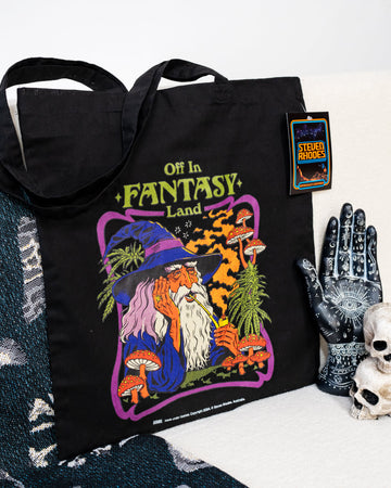 Steven Rhodes black tote bag featuring a whimsical design titled ‘Off in Fantasy Land.’ The artwork depicts a wizard sipping a drink surrounded by mushrooms and magical plants, with bold purple, orange, and green tones. The bag includes sturdy handles and an eco-friendly label, combining functionality with quirky charm.