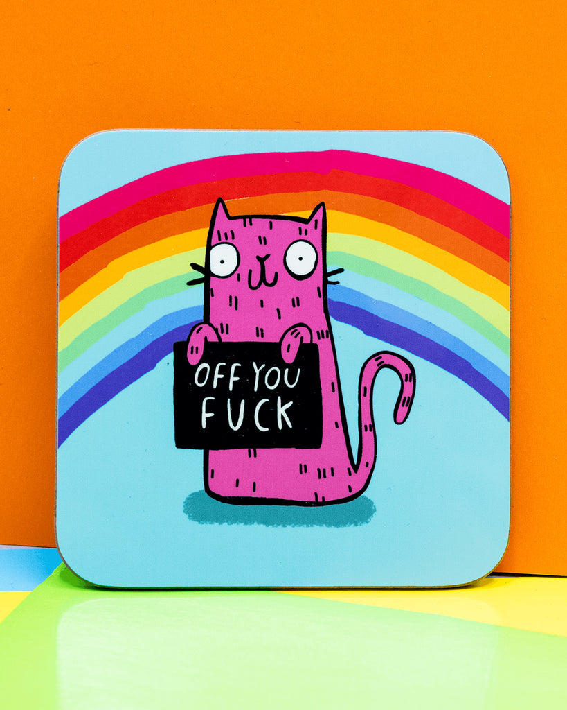 Colourful square coaster featuring a pink cartoon cat holding a black sign that says “Off You F*ck,” set against a light blue background with a rainbow arch. Designed by Katie Abey.