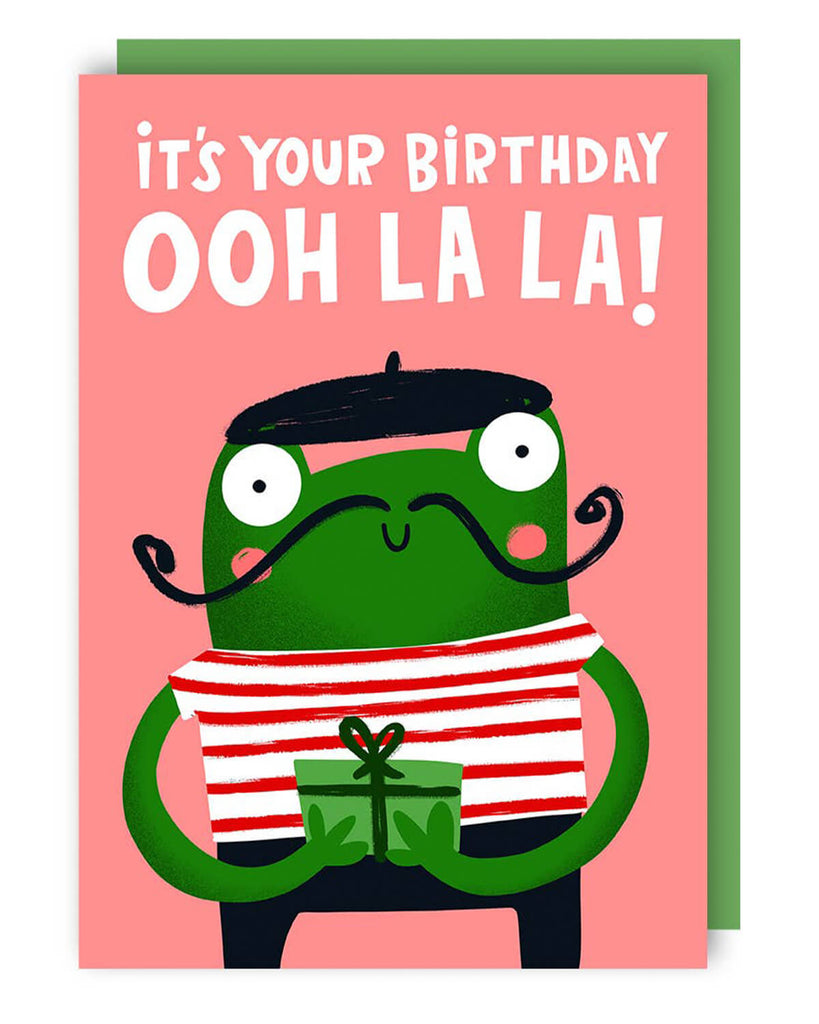 Quirky birthday card from Rebel Romance featuring a green cartoon frog in a red striped shirt and beret, with a curly moustache and a gift box. Text reads “It’s your birthday – ooh la la!” in bold white letters on a pink background. A fun and playful greeting card perfect for alternative birthday wishes with a touch of French flair and frog-themed charm.