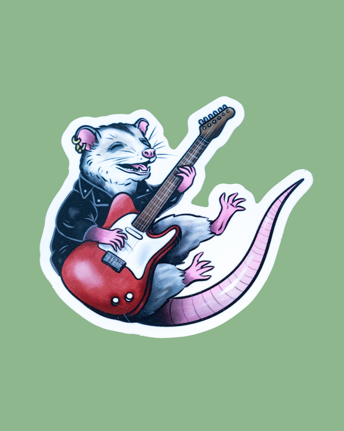 Vinyl sticker of a grinning opossum wearing a black leather jacket and gold earring, playing a red electric guitar. The opossum has pink paws, a curled tail, and joyful expression. Design by Rebel Romance, set against a pale background with dimensions 7.2 cm x 6.7 cm shown.