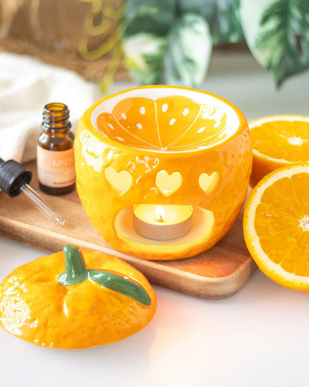 The orange oil burner in action, with a lit tealight candle inside creating a warm glow. The heart-shaped cutouts cast soft, flickering light, enhancing the cosy atmosphere. The fragrance dish is filled with oil, ready to diffuse a refreshing scent.