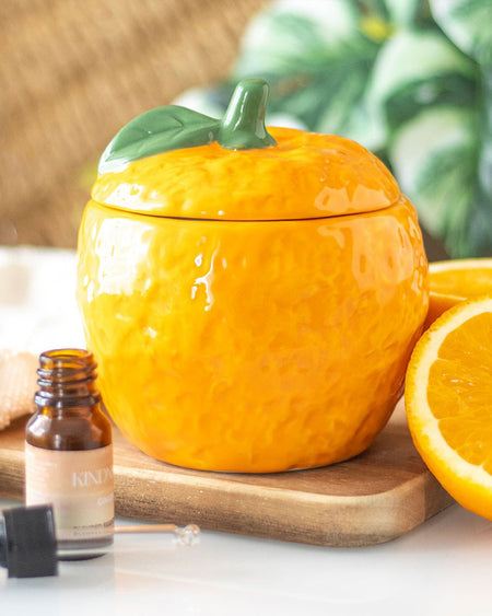 A beautifully styled image featuring the orange oil burner placed on a wooden surface next to an essential oil dropper bottle and fresh orange slices. The bright citrus tones and realistic design make it a fun and quirky decorative piece for kitchens, bedrooms, or living spaces.