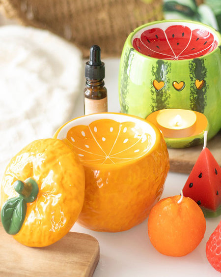 A side-by-side styling of the orange oil burner and a watermelon-themed oil burner, both placed on a bright and cheerful setup. Small fruit-shaped candles, essential oils, and fresh fruit slices add to the aesthetic, showcasing the playful, retro-inspired homeware collection by Rebel Romance.