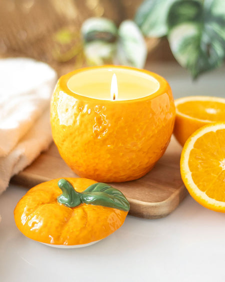 The orange candle jar lit, creating a warm and cosy glow. The ceramic lid rests beside it, showcasing its detailed fruit-inspired design.