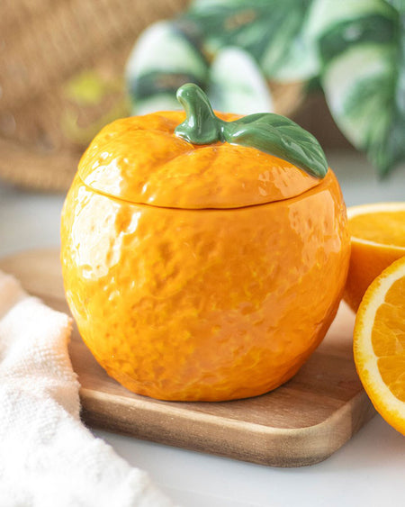 The orange-shaped candle jar styled on a wooden surface, surrounded by fresh orange slices. Its realistic citrus design adds a pop of colour to any room.