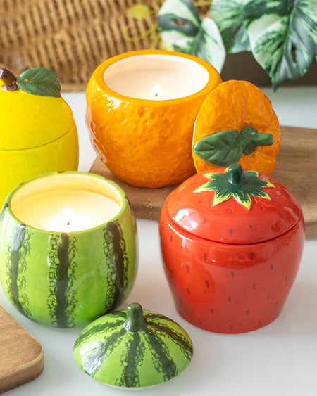 A collection of fruit-shaped candle jars, including orange, lemon, strawberry, and watermelon designs. The ceramic jars feature removable lids and textured surfaces, creating a vibrant and playful home décor aesthetic.