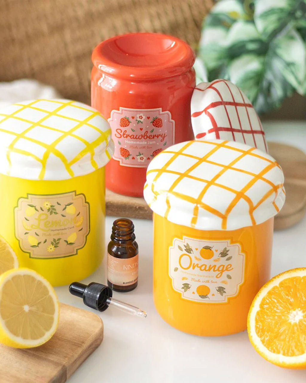A collection of ceramic jam jar oil burners in lemon, orange, and strawberry designs. The lids mimic traditional gingham-patterned jam jar covers. The orange marmalade burner is in focus, placed among fresh citrus slices and an essential oil bottle.