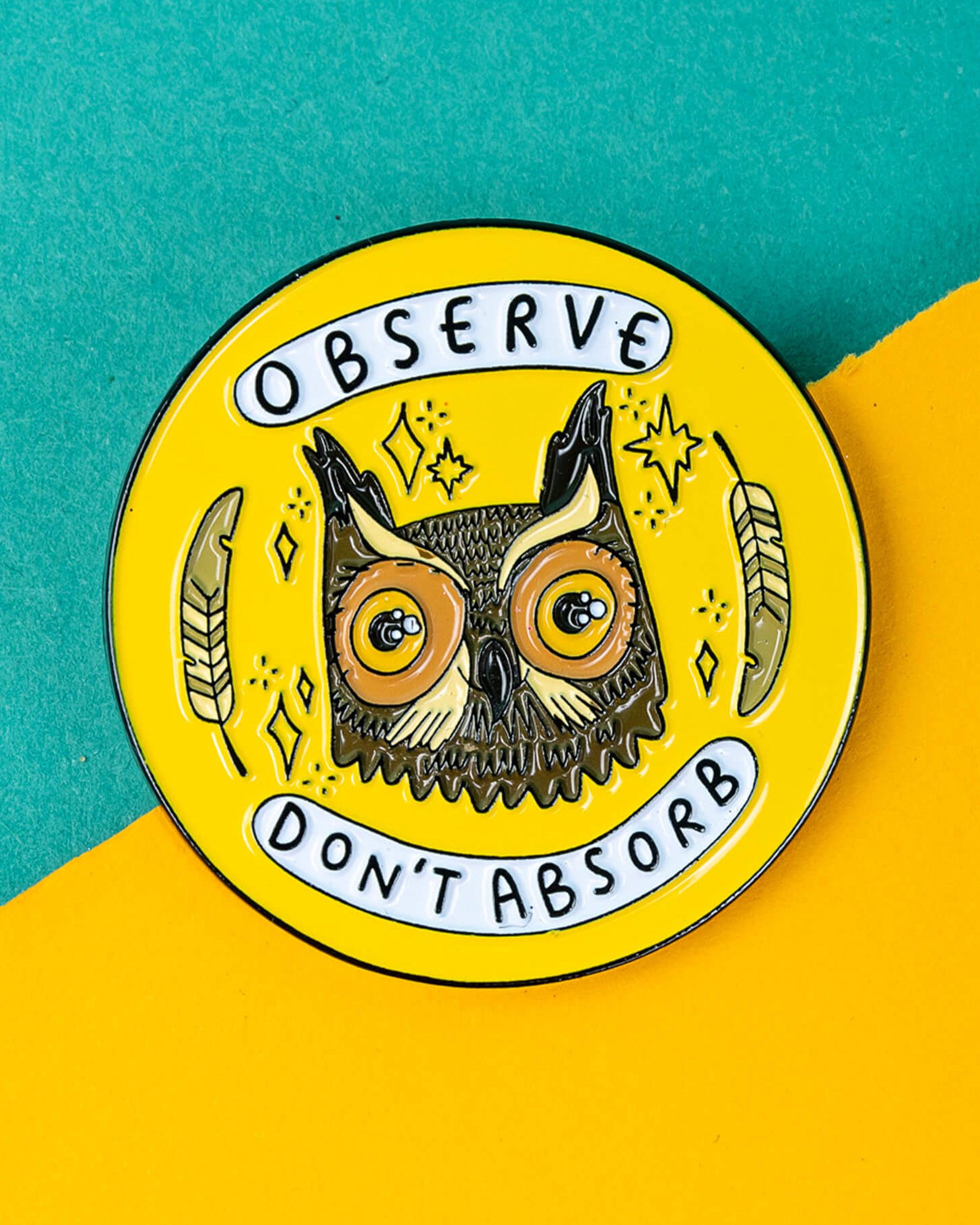 Close-up of the Rebel Romance “Observe Don’t Absorb” owl enamel pin by Katie Abey on a split teal and yellow background. The round pin shows a wise owl with feathers and stars, with bold text reading “Observe” and “Don’t Absorb.” A bright, meaningful accessory loved by fans of alternative fashion and unique gifts.