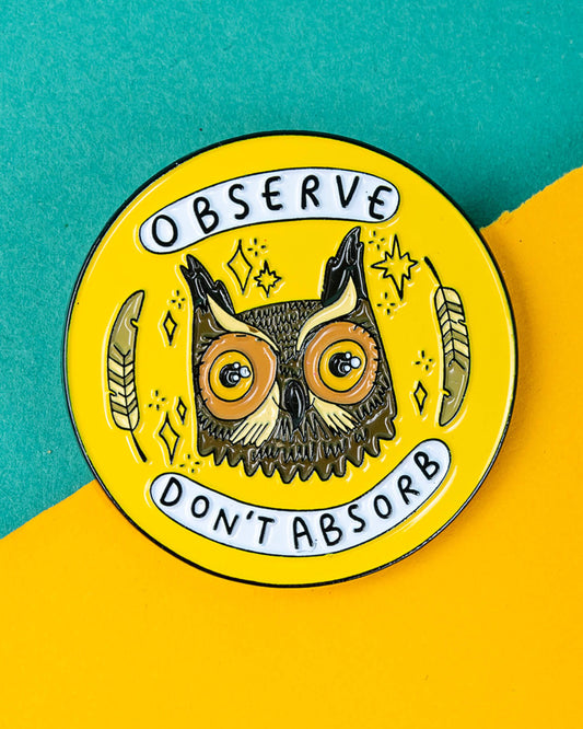 Close-up of the Rebel Romance “Observe Don’t Absorb” owl enamel pin by Katie Abey on a split teal and yellow background. The round pin shows a wise owl with feathers and stars, with bold text reading “Observe” and “Don’t Absorb.” A bright, meaningful accessory loved by fans of alternative fashion and unique gifts.