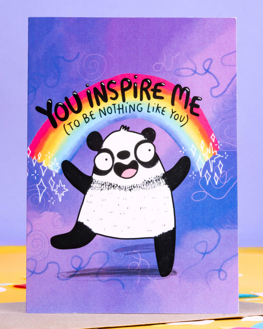 A colourful A6 greeting card by Rebel Romance featuring a cheeky rainbow panda illustration by Katie Abey. The card reads “You Inspire Me (To Be Nothing Like You)” in bold letters above a happy panda under a rainbow, on a vibrant purple background with doodle-style stars and swirls. Ideal for sending playful, sarcastic messages to friends or frenemies. Blank inside for your own note.