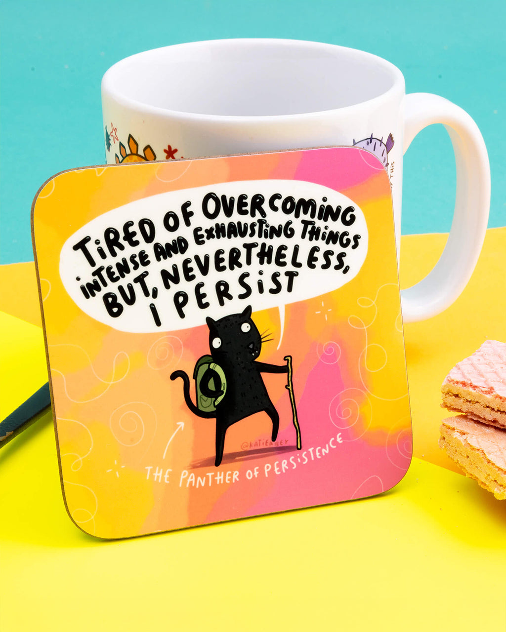 A square coaster with rounded corners featuring a cartoon black panther with a backpack and walking stick, illustrated by Katie Abey. Text reads: “Tired of overcoming intense and exhausting things but, nevertheless, I persist.” Set against a warm orange-pink gradient. A motivational coaster for hot drinks, perfect for desks or workspaces.