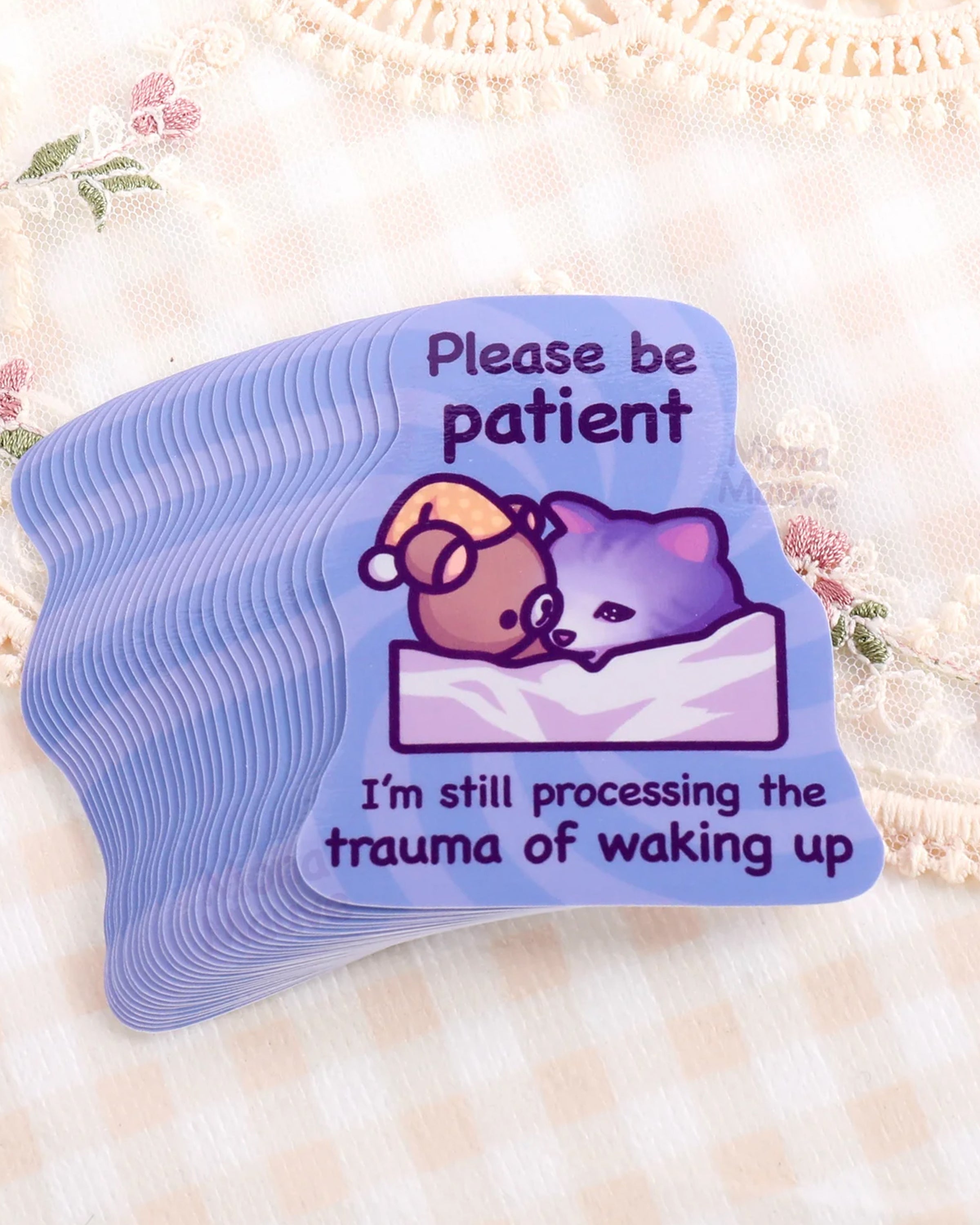 Stack of Rebel Romance funny cat meme vinyl stickers by Mona Mauve, featuring a sleepy purple cat in bed with a teddy bear and the text “Please be patient, I’m still processing the trauma of waking up.” Printed on a pastel purple background with waterproof, durable vinyl. Perfect for laptops, journals, or gifting to fellow night owls. Set against vintage floral lace fabric. A relatable and humorous sticker for tired millennials and alternative stationery lovers.