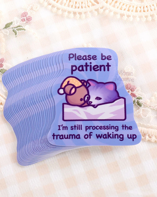 Stack of Rebel Romance funny cat meme vinyl stickers by Mona Mauve, featuring a sleepy purple cat in bed with a teddy bear and the text “Please be patient, I’m still processing the trauma of waking up.” Printed on a pastel purple background with waterproof, durable vinyl. Perfect for laptops, journals, or gifting to fellow night owls. Set against vintage floral lace fabric. A relatable and humorous sticker for tired millennials and alternative stationery lovers.