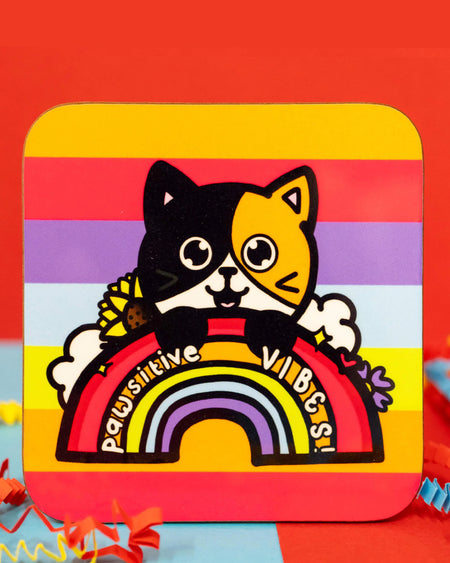 Close-up of the Pawsitive Vibes Cat Coaster by Innabox. The square coaster has rounded corners and a vibrant rainbow-striped background. The design features an adorable calico cat with one black side and one orange side, peeking over a bold red rainbow with the words "Pawsitive Vibes!" written across it. Sunflowers, lavender, and fluffy clouds add extra charm. Colourful crinkle paper surrounds the coaster. Available on Rebel Romance